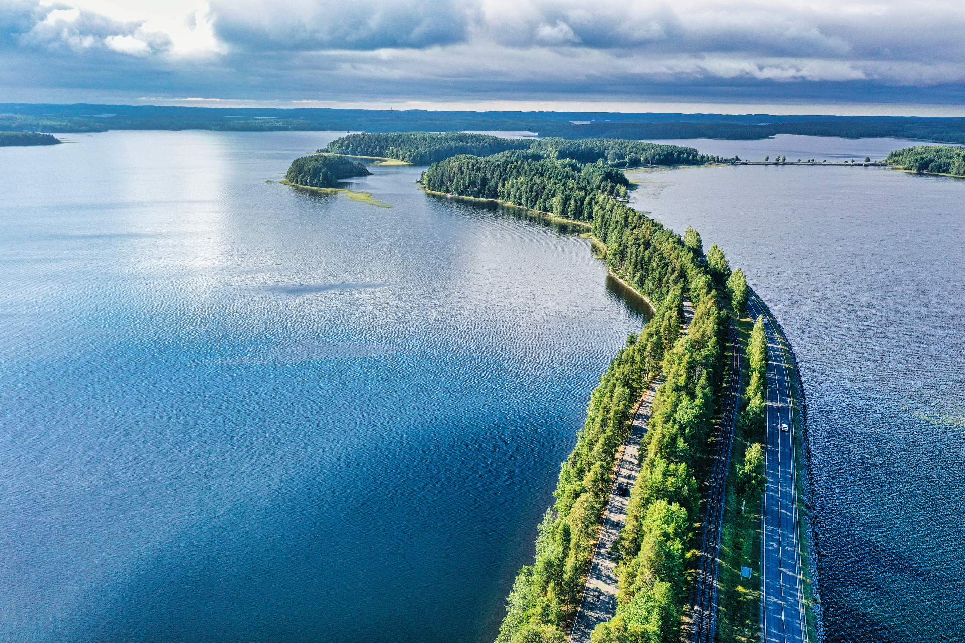 Ilmakuva järvestä, metsäsaarista ja pitkistä tie- ja rautatiesilloista, pilvinen taivas yllä.