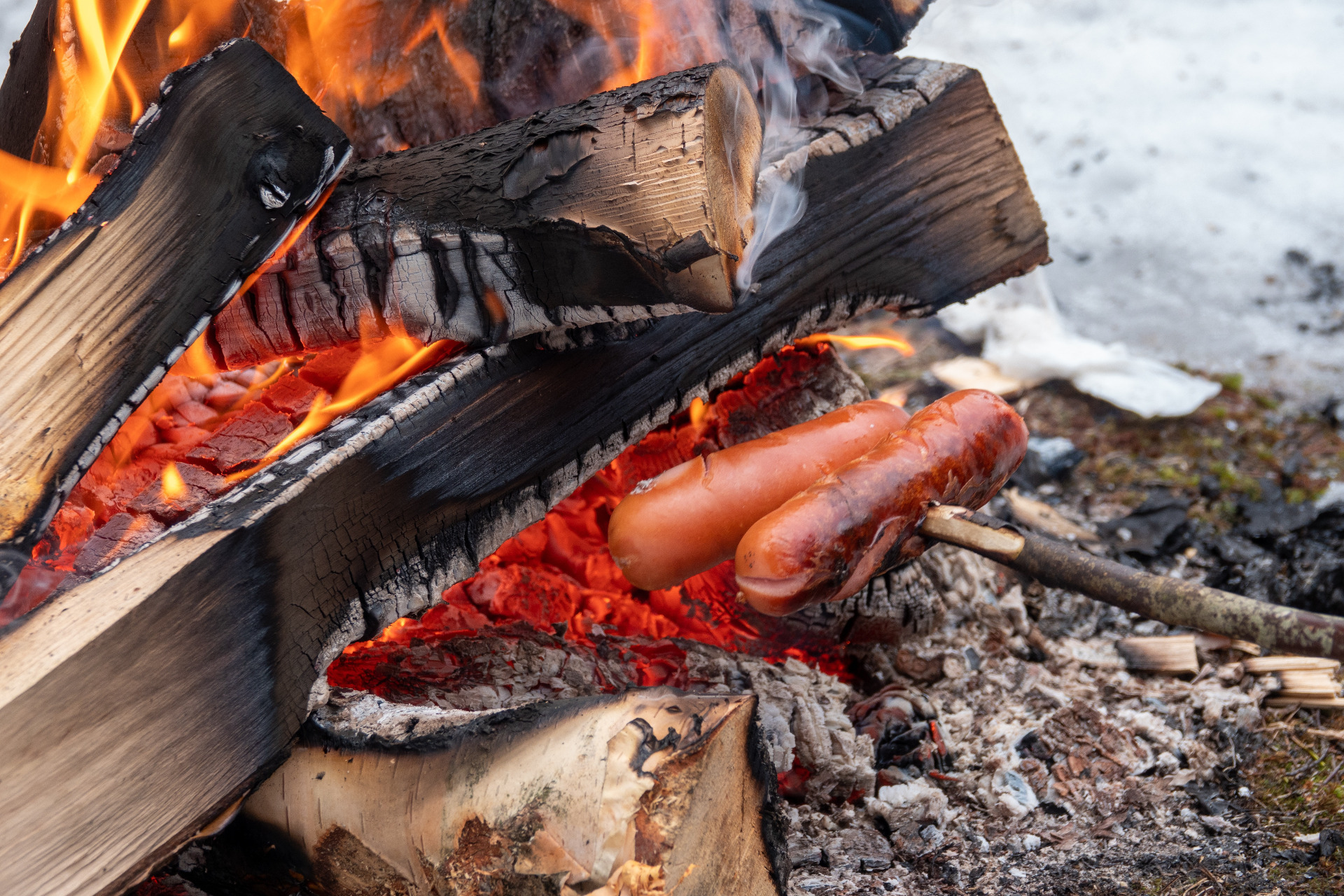 Avotulen hiilloksella grillatut makkarat ja puunloki, liekit ja savua näkyvät taustalla.