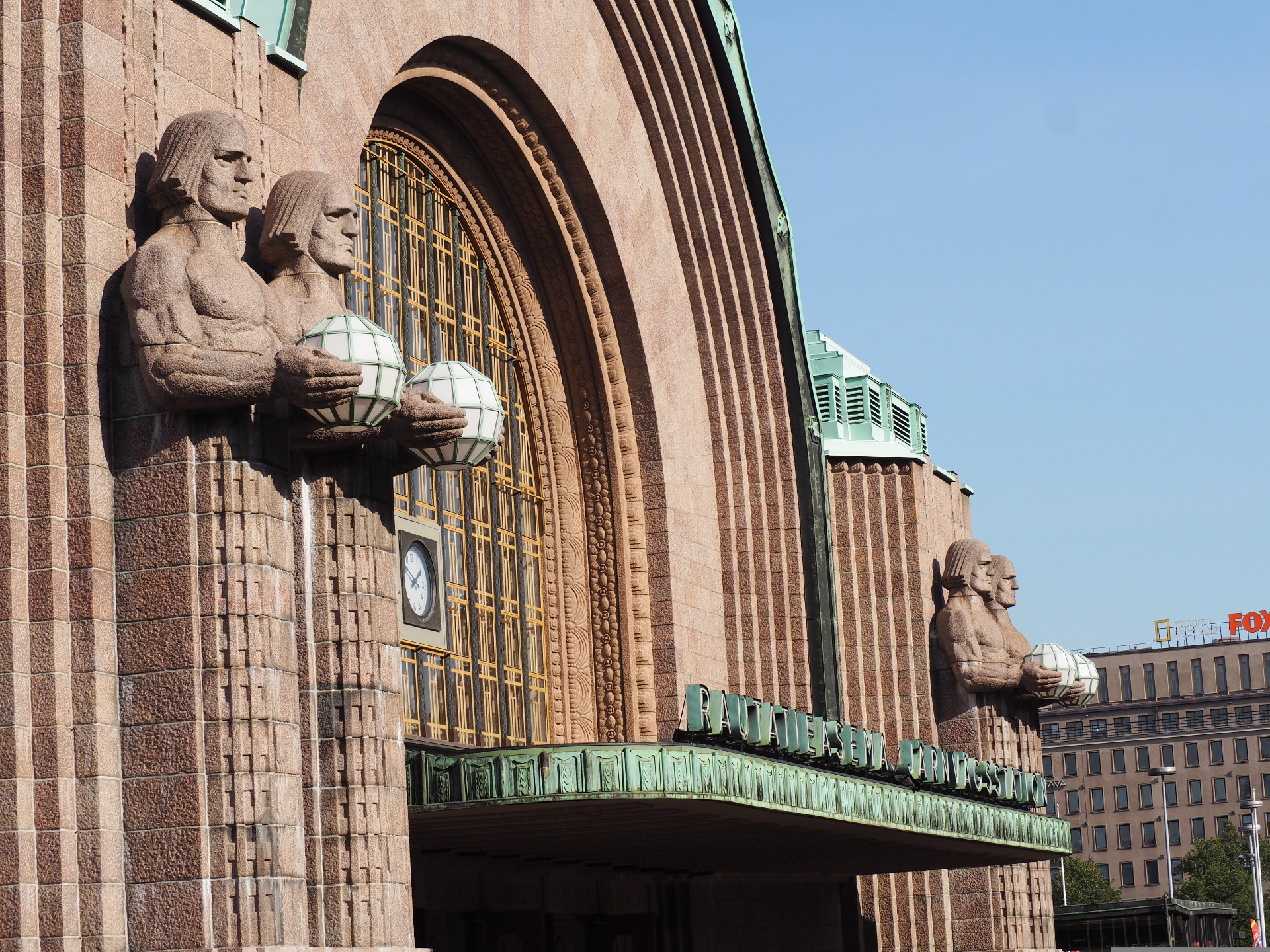 Kivijulkisivuun veistetyt patsaat ja suuret lyhdyt, taustalla kaareva ikkuna ja sininen taivas. Helsingin rautatieasema.