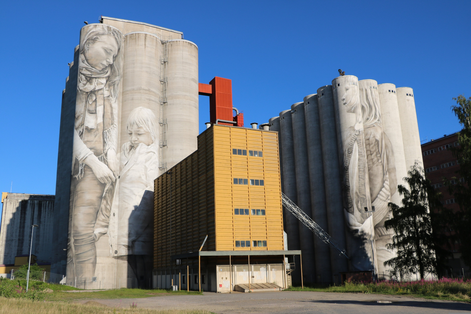 Teollisuuslaitoksen korkeat siilot, joissa on valkoiset veistokselliset kasvokuvapinnat, sininen taivas ja keltainen rakennus. Kantolan muraali, Hämeenlinna.