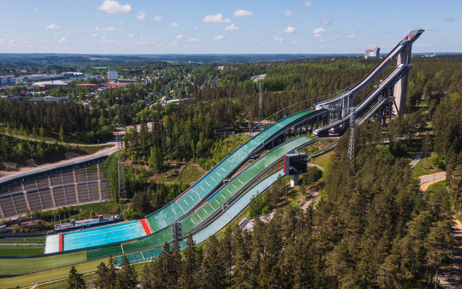 Ilmakuva mäkihyppyrimäestä, vihreästä radasta ja kaupunkimaisemasta metsän keskellä, sininen taivas pilvien kanssa. Salpauselkä, Lahti matkailu