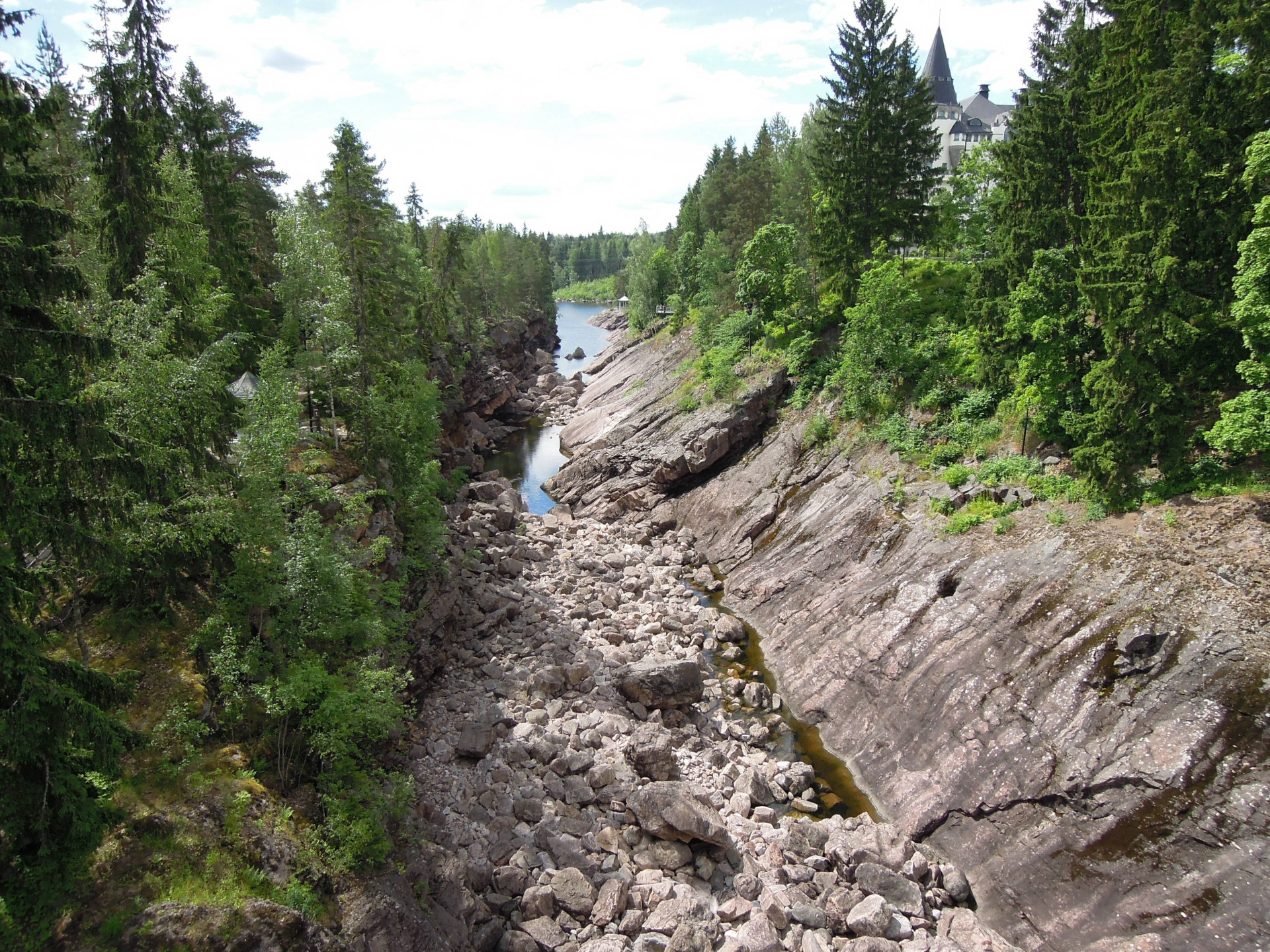 Kapea joki virtaa kivikkoisessa uomassa tiheässä metsässä, taustalla linna tornineen. Imatrankoski. Imatra matkailu