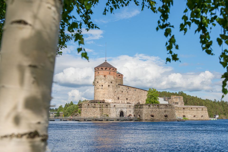 Kivinen linnoitus järven rannalla, punakattoinen torni ja lehtiä kehyksenä sinisellä taivaalla. Olavinlinna. Savonlinna matkailu.