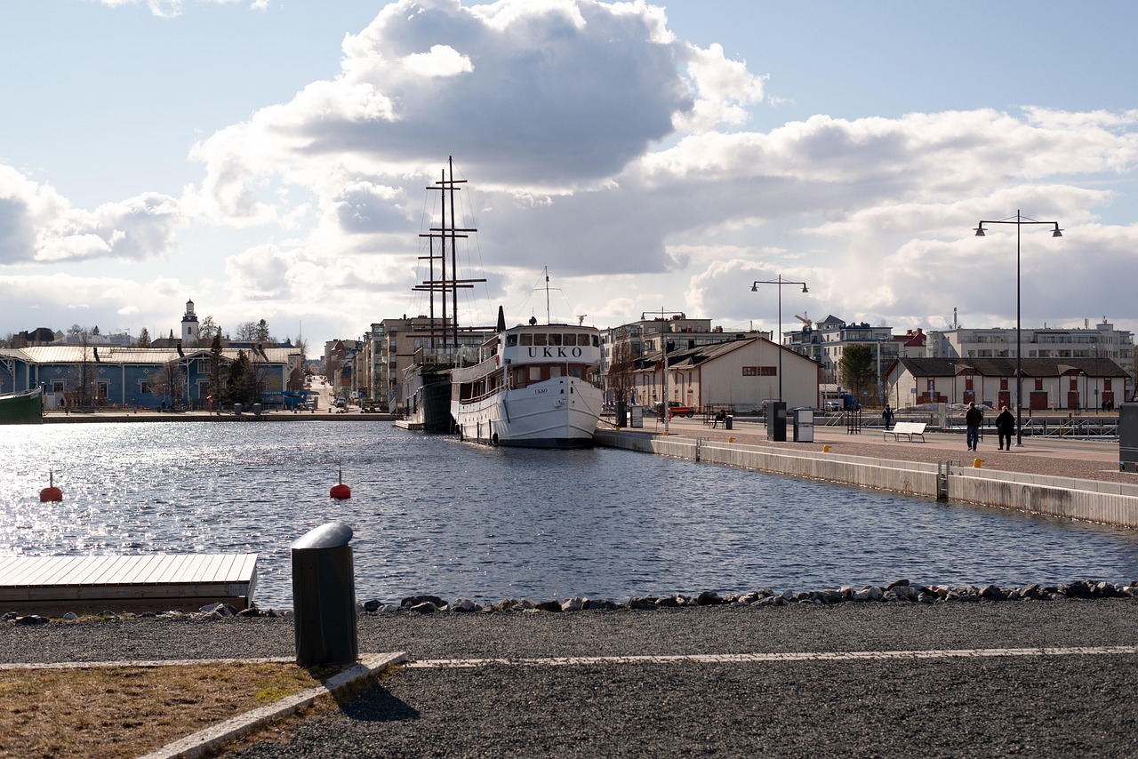 Satamakuva, purjealus “UKKO” laiturissa, pilvinen taivas ja kaupunkirakennuksia joen rannalla. Kuopion satama, Kuopio matkailu.