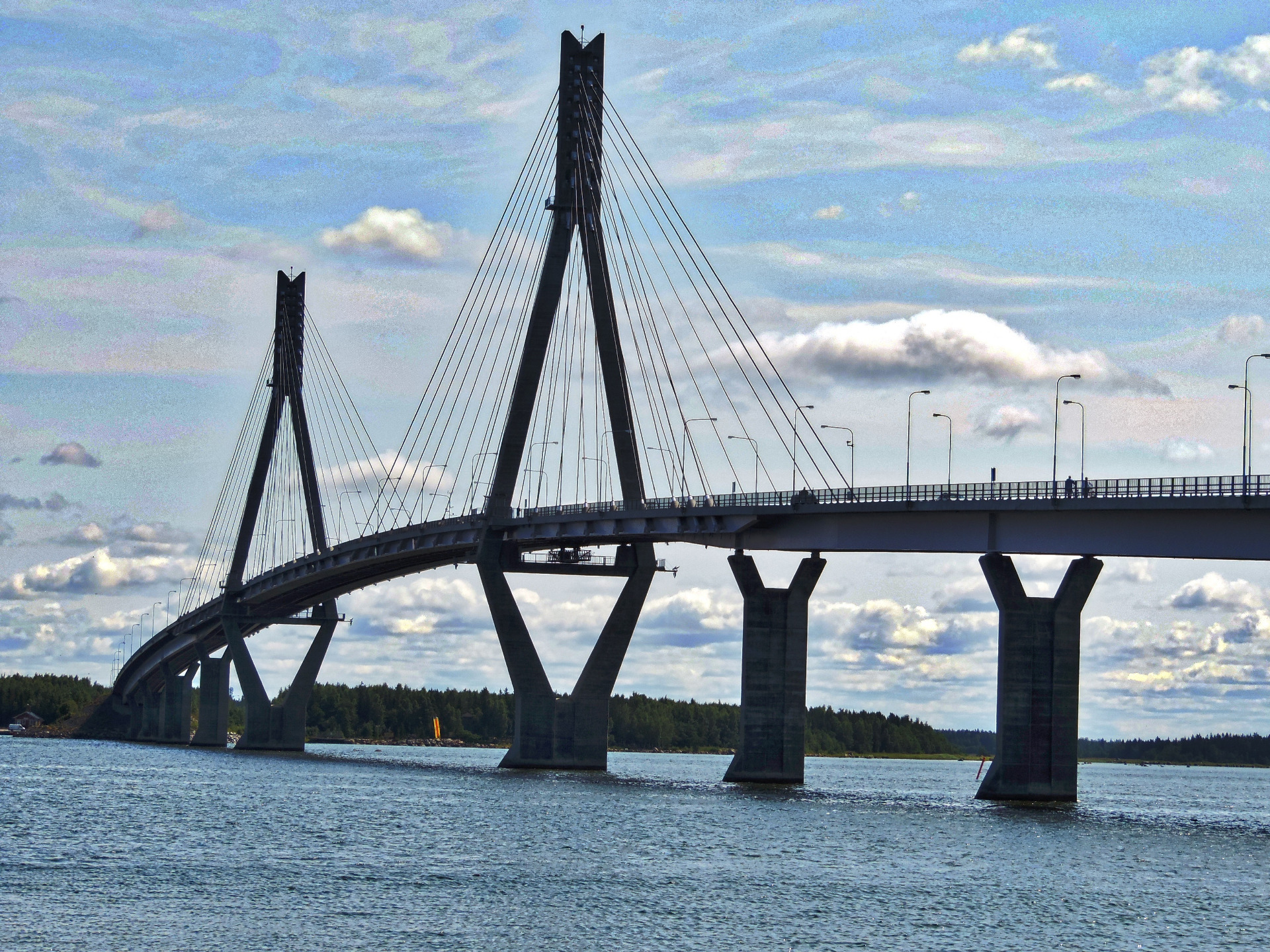 Jousisilta veden yllä, korkeat tornit ja vinot kaapelit, taustalla pilvinen taivas ja metsäranta. Raippaluoto, Vaasa, Mustasaari. Matkailu.