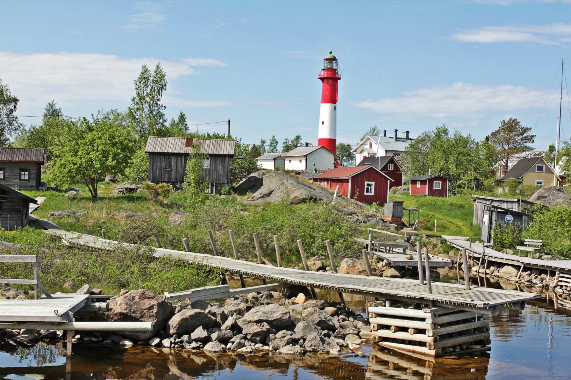 Punavalkoinen majakka kohoaa rantakylän yllä, puutaloja ja laiturit, taustalla sininen taivas ja pilviä. Tankar. Majakka. Kokkola