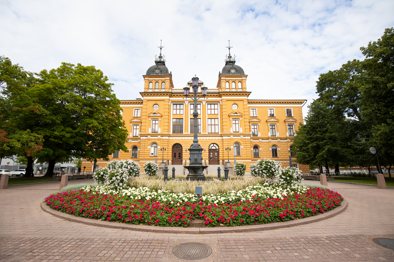 Keltainen historiallinen rakennus, keskellä suihkulähde ja kukkapenkki, ympärillä vihreitä puita ja pilvinen taivas. Oulu matkailu. Oulun kaupungintalo.