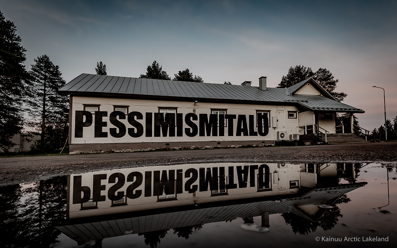 Vanha rakennus, jossa teksti “PESSIMISMITALO” ja peilikuva lammikossa, taustalla metsää ja pilvinen taivas. Puolanka matkailu. Puolanka.