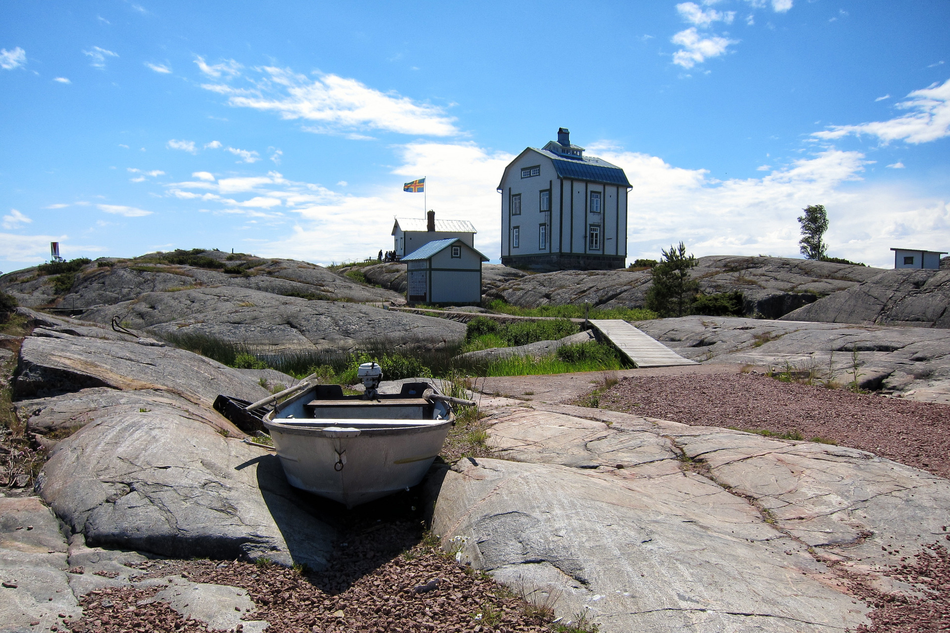 Valkoinen vene kivikkoisella rannalla, taustalla sininen talo ja lipputanko kirkkaalla taivaalla. Ahvenanmaan saaristo. Åland. Visit Maarinahamina.
