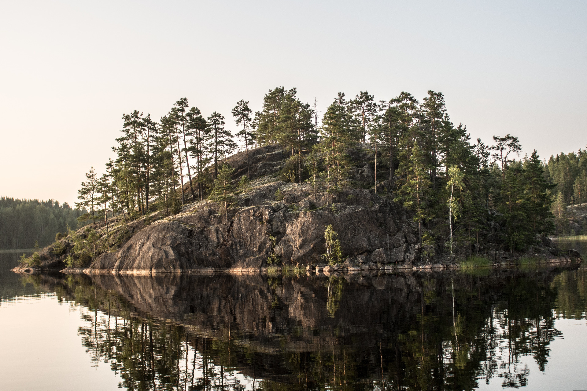 Metsäinen kivikkorinne järven rannalla, puiden heijastukset näkyvät tyynessä vedessä. Saimaa. Unesco Global Geopark.