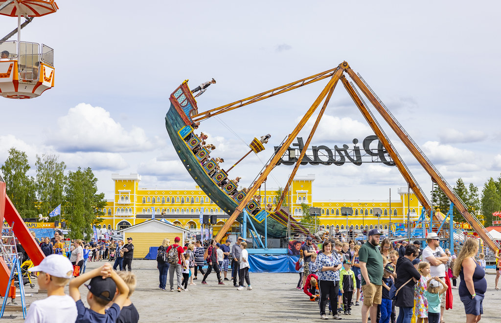 Juhlavieraita huvipuistossa, suuri keinurakenne ja taustalla keltainen rakennus pilvisellä säällä. Miljoonativoli. Tivoli Sariola. Tuuri. Keskisen Kyläkauppa.