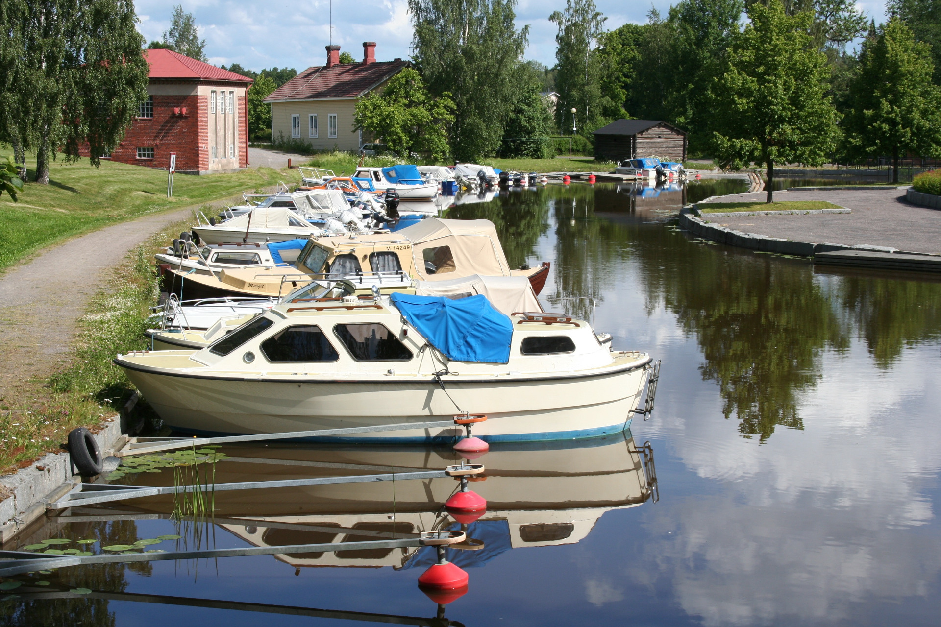 Valkoisia ja sinisiä veneitä laiturissa rauhallisella kanavalla, taustalla punatiilinen rakennus ja vehreää puistoa. Forssa matkailu. Forssan kansallinen kaupunkipuisto.