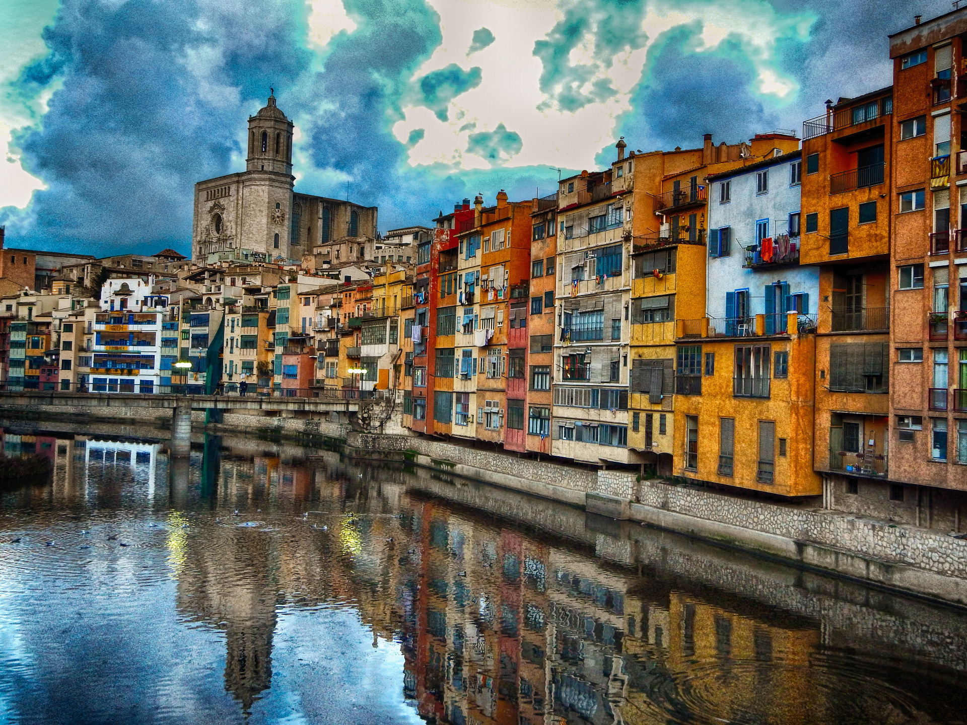 Värikkäitä kerrostaloja joen rannalla, taustalla kirkontorni ja pilvinen taivas heijastuu veteen. Espanja. Catalonia - Girona.