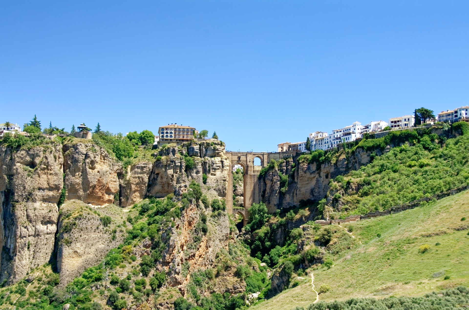 Kallioinen laakso, vihreää kasvillisuutta ja kivisilta, taustalla valkoisia rakennuksia sinisellä taivaalla. Espanja. Ronda. Andalucia