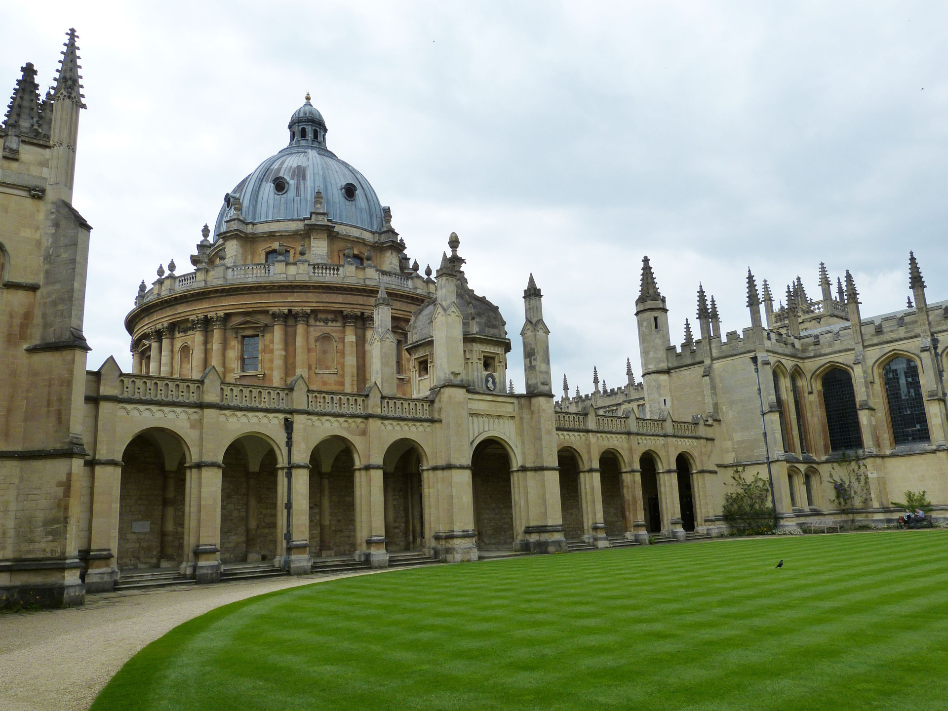 Kirkon kupoli ja kivikaaret, taustalla goottilaisia torneja, nurmikkoa ja harmaa taivas. Oxford on idyllinen opiskelijakaupunki.