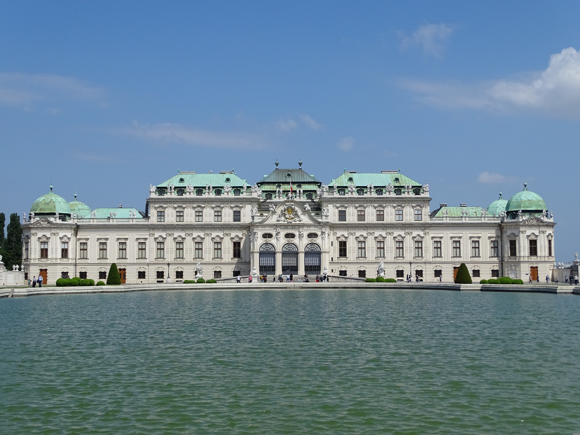 Historiallinen rakennus vihreine kuparikattoineen ja kupoleineen, edustalla vettä ja kävelijöitä sinisellä taivaalla. Wien. Itävalta. Belvederen linna.
