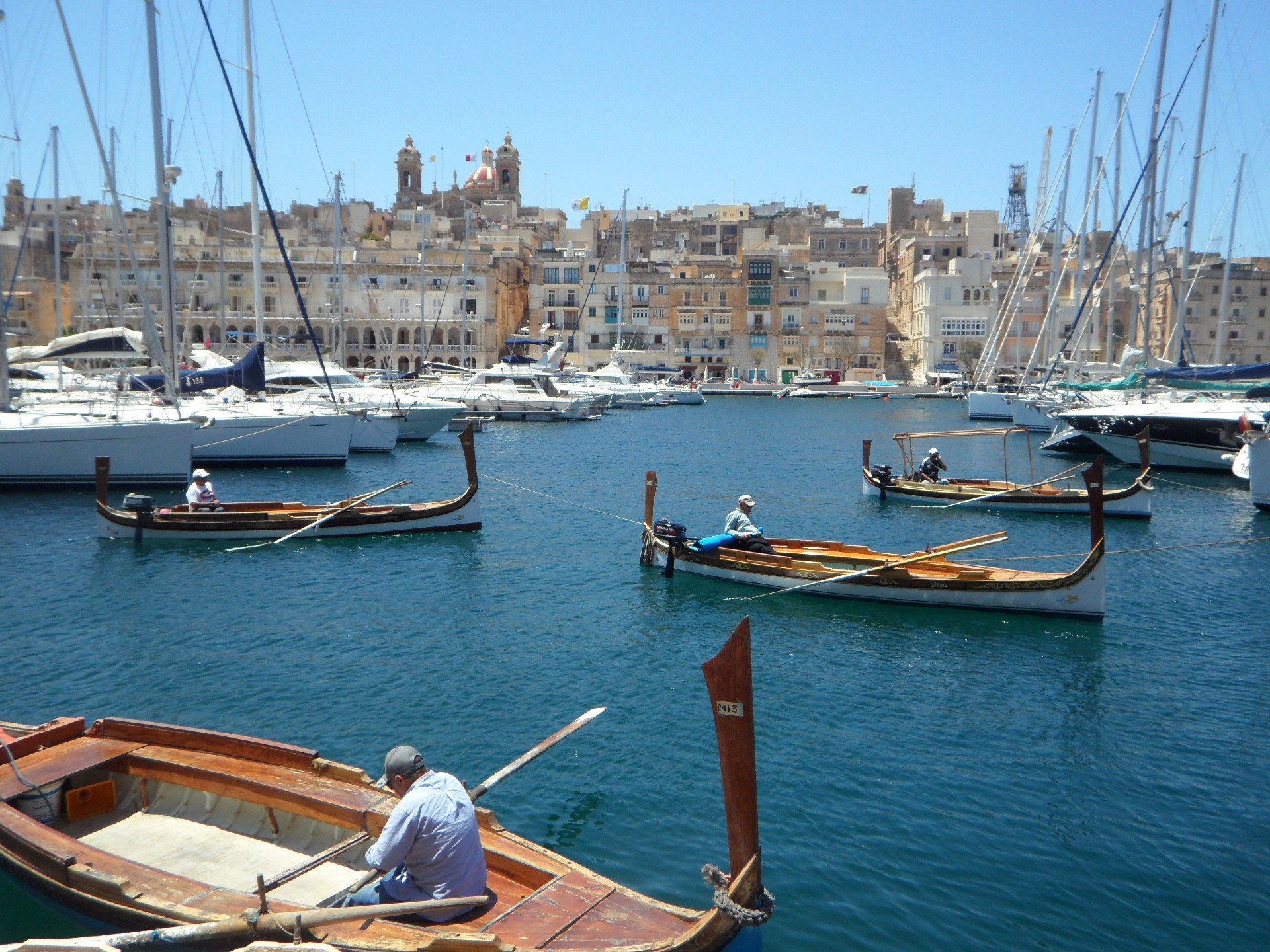 Maltan Valletta. Malta on kaunis saarivaltio Välimerellä. Purjeveneitä satamassa, taustalla tiheä kaupunkimaisema ja kirkontornit sinisellä taivaalla.