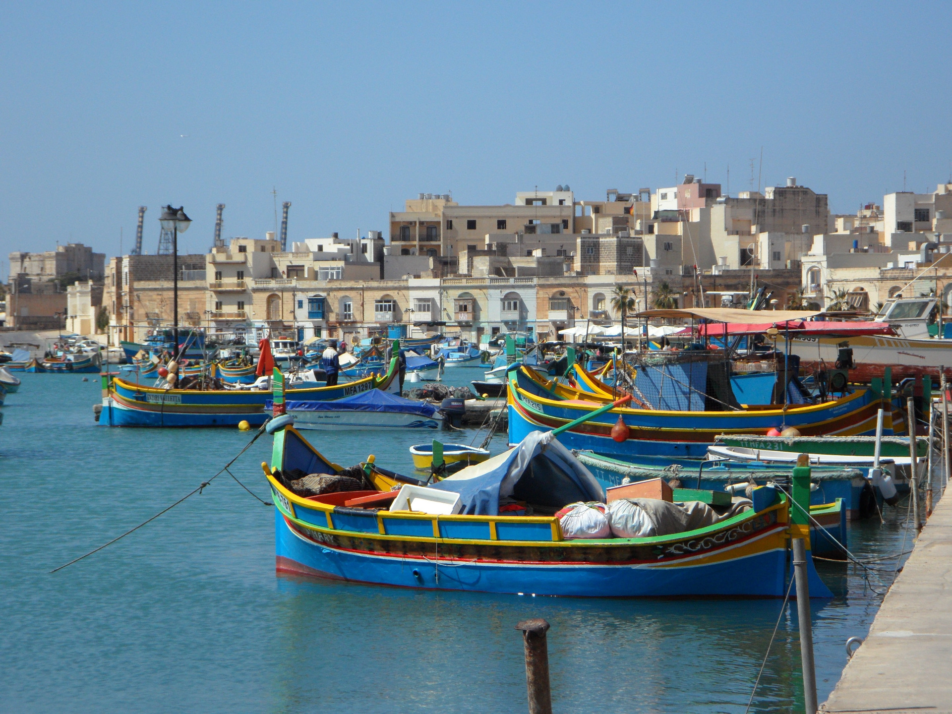 Marsaxlokk. Malta. Sininen satama täynnä kalastusaluksia, taustalla tiiviisti rakennettu kaupunkimaisema ja kirkas taivas.