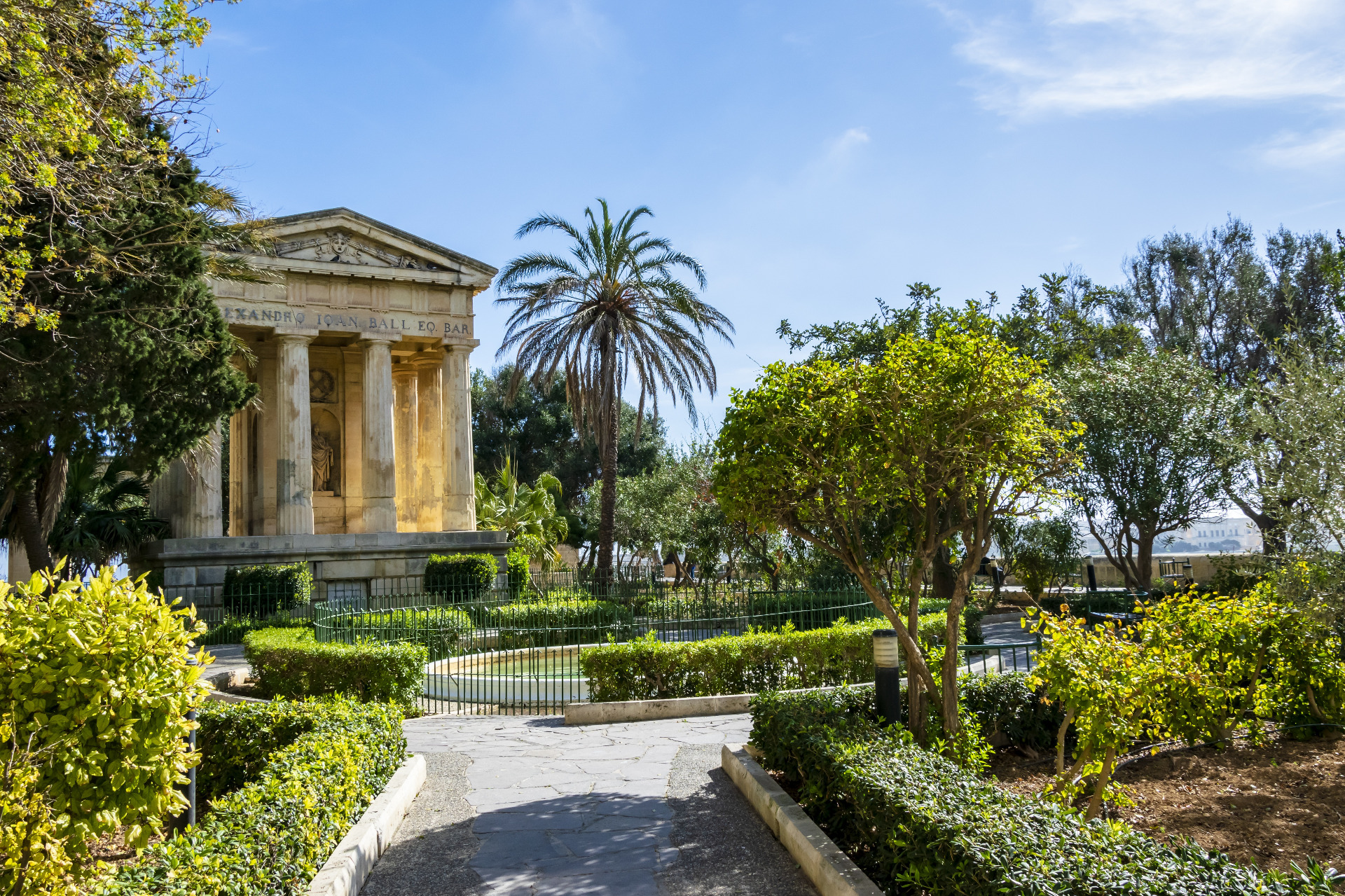 Malta. Valletta on historiallisia rakennuksia. Klassinen kivitemppeli pylväineen, palmuja ja vehreää puutarhaa sinistä taivasta vasten.