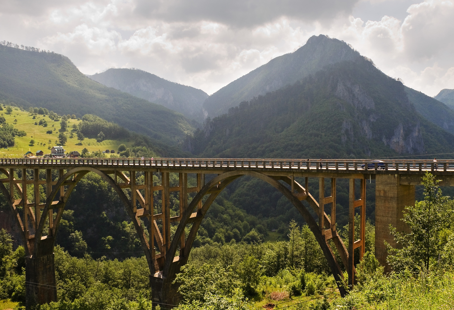 Puinen rautatiesilta kaartaa vuorilaakson yli, taustalla vihreitä rinteitä ja pilvinen vuorenhuippu. Montenegro, Djurdjevicin silta, Tara Kanjoni