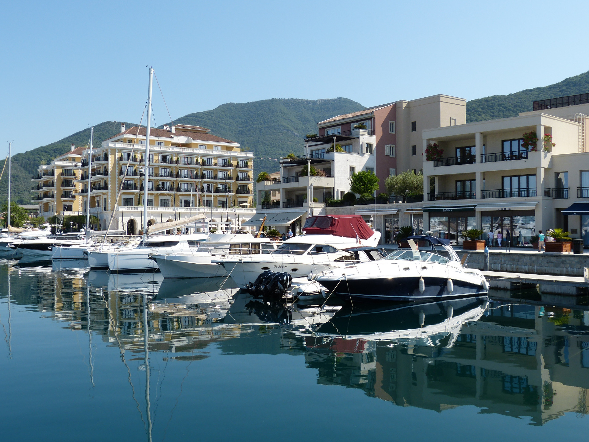 Valkoisia huvialuksia satamassa, taustalla hotellirakennuksia ja vihreä vuorimaisema sinisellä taivaalla. Kotorin satama on vilkas, Montenegrossa.
