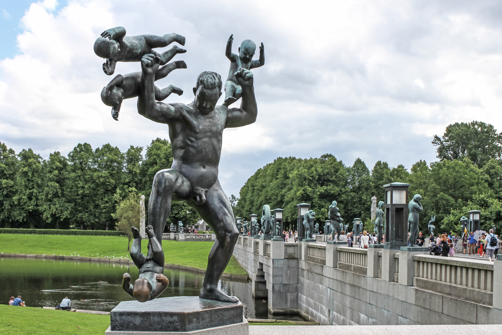 Vigeland park, Oslo. Norja. Pronssinen veistosryhmä nostaa käsiä ja jalkoja puistossa, taustalla lampi, ihmiset ja pilvinen taivas.