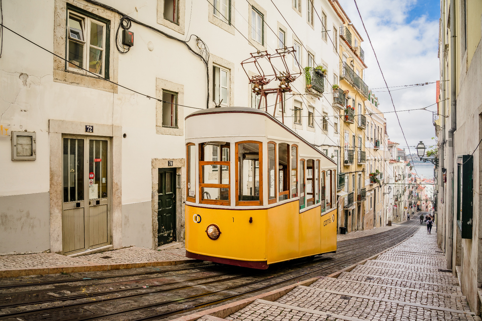 Lissabon Portugali. Kauniit raitiovaunut kuuluvat katukuvaan. Keltainen raitiovaunu kulkee kapealla mukulakivikadulla, vanhojen rakennusten ja sähköjohtojen ympäröimänä.