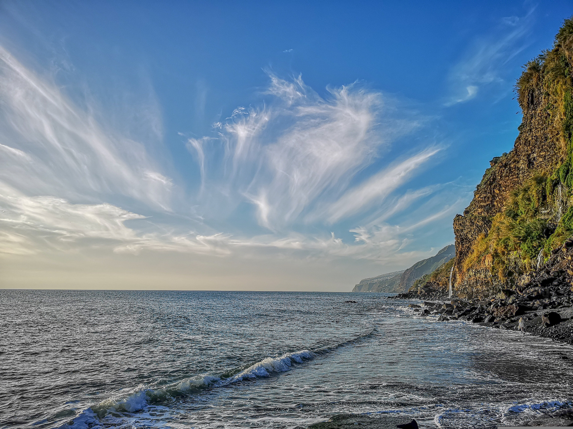 Luonnonkaunis Madeira on suosittu lomakohde, Portugalissa. Sininen taivas ja aaltoileva meri, kallioinen rantaviiva oikealla, pilvijuovat ja auringonvalo.