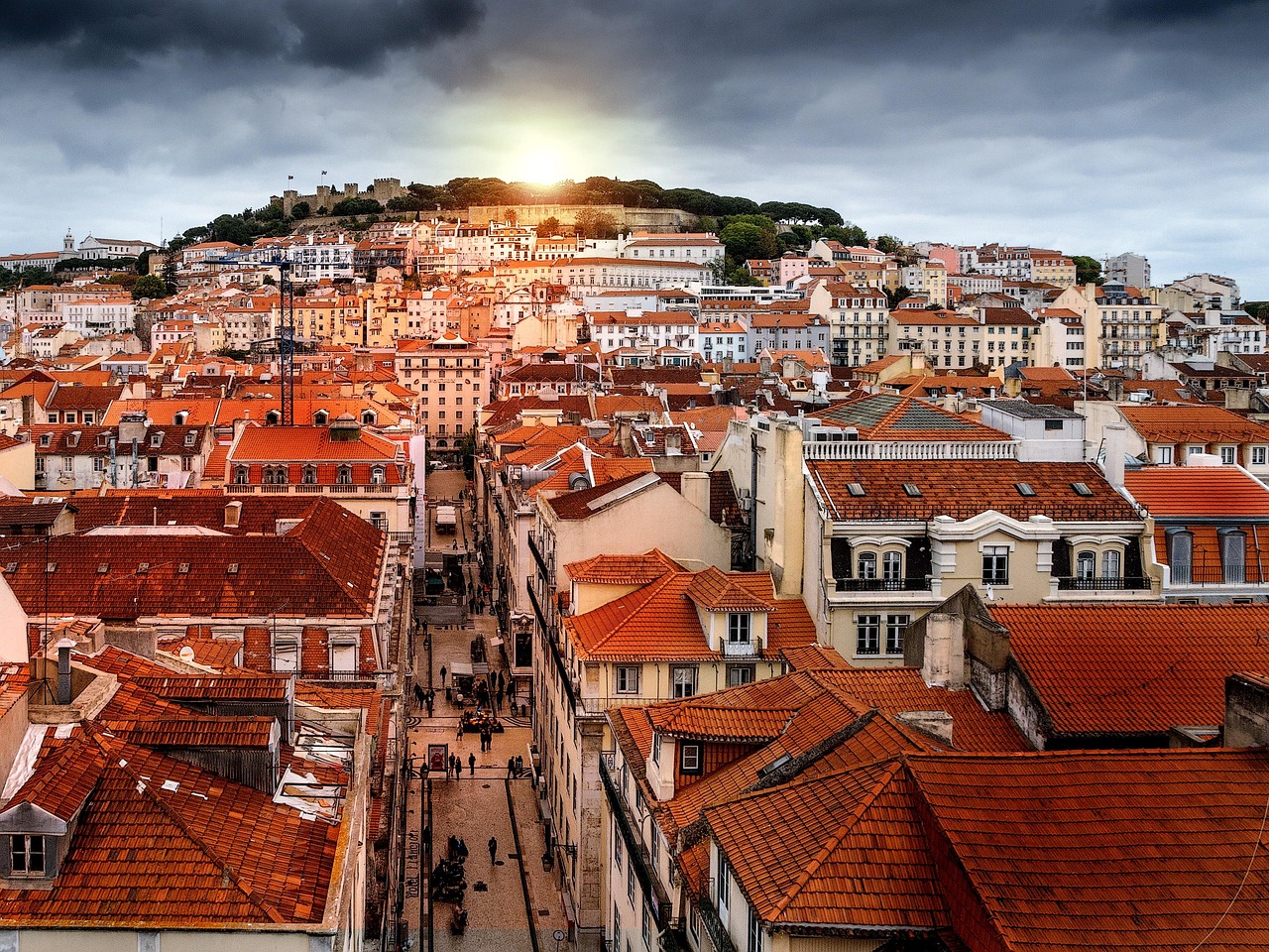 Lissabon Portugalissa on eläväinen matkailukaupunki. Punatiilikattoisia rakennuksia ja kapeita katuja kukkulan rinteessä, auringonlasku pilvien läpi.