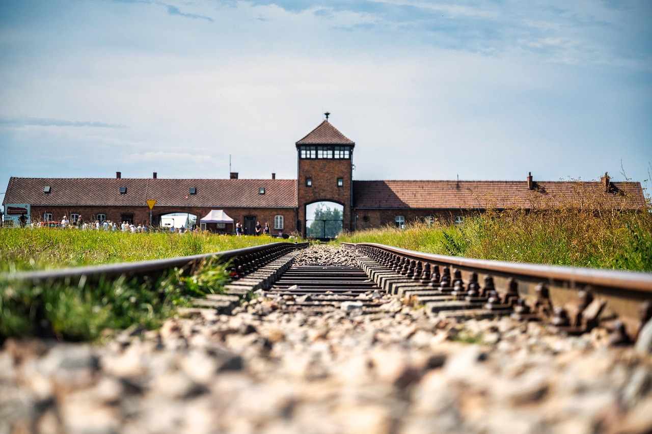 Auschwitz I ja Birkenau ovat tunteita herättäviä matkakohteita Puolassa. Rautatien kiskot johtavat kohti tiilirakennusta, taustalla ihmisjoukko ja sininen taivas pilvien lomassa.