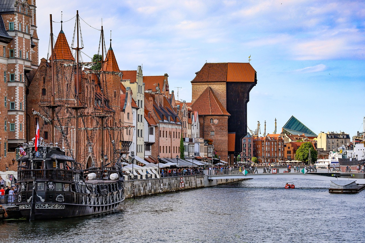 Gdansk, Puola on eläväinen Hansakaupunki. Punatiilisiä vanhoja rakennuksia ja kanava, purjelaivamainen alus, ihmisjoukkoja ja punainen kattoinen torni taustalla.
