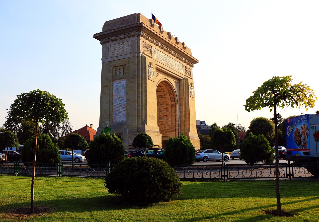 Bucharest, Arc de triomphe riemukaari on vakuuttava nähtävyys. Monumentaalinen kivikaari, liput ylhäällä, puisto ja pensaita sekä pysäköityjä autoja taustalla.