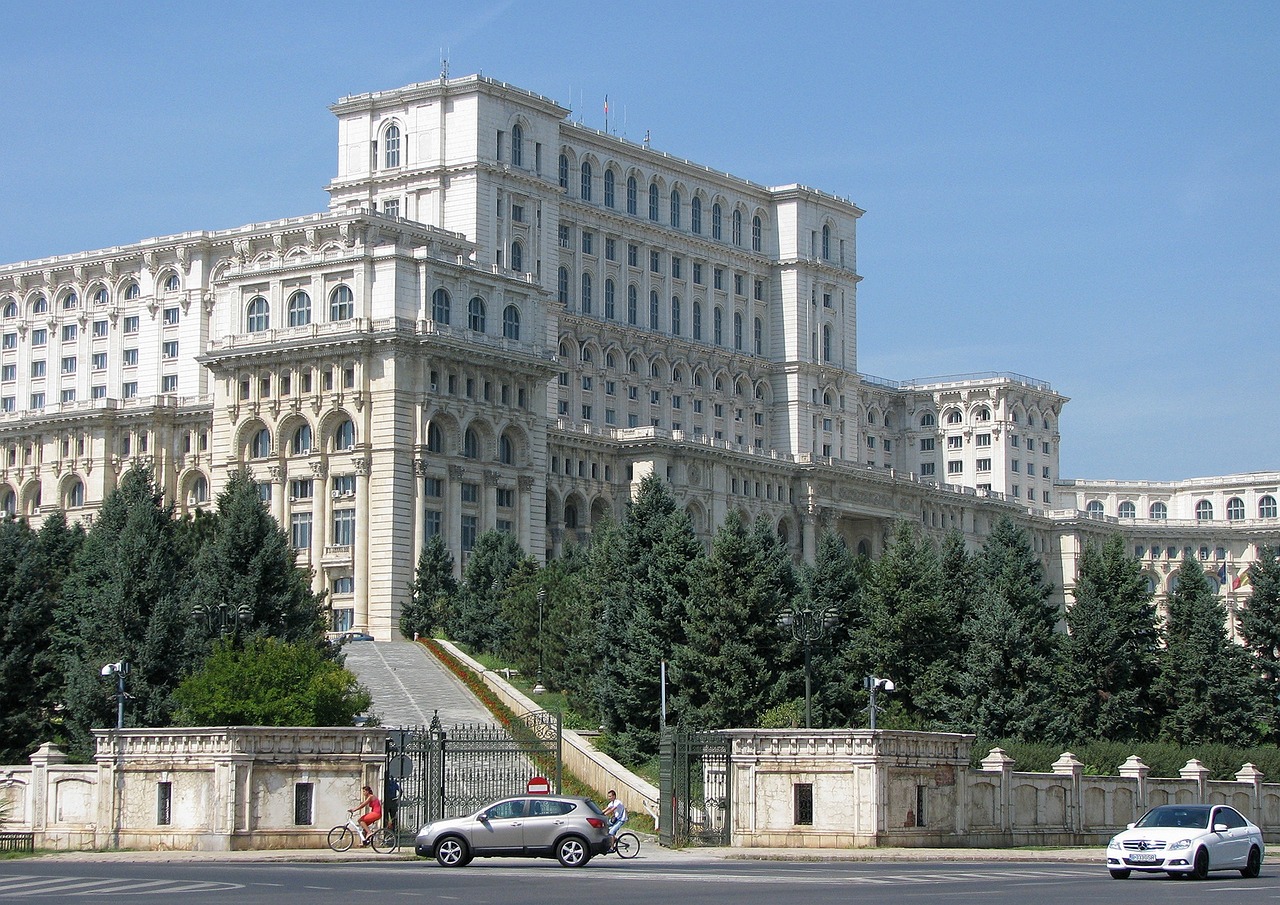 Kansantalo, Parlamenttitalo, Nicolae Ceaușescu palatsi, Bukarest. Valkoinen monumentaalinen rakennus, kaarevia ikkunoita ja pylväitä, edessä puita, autot ja pyöräilijöitä.