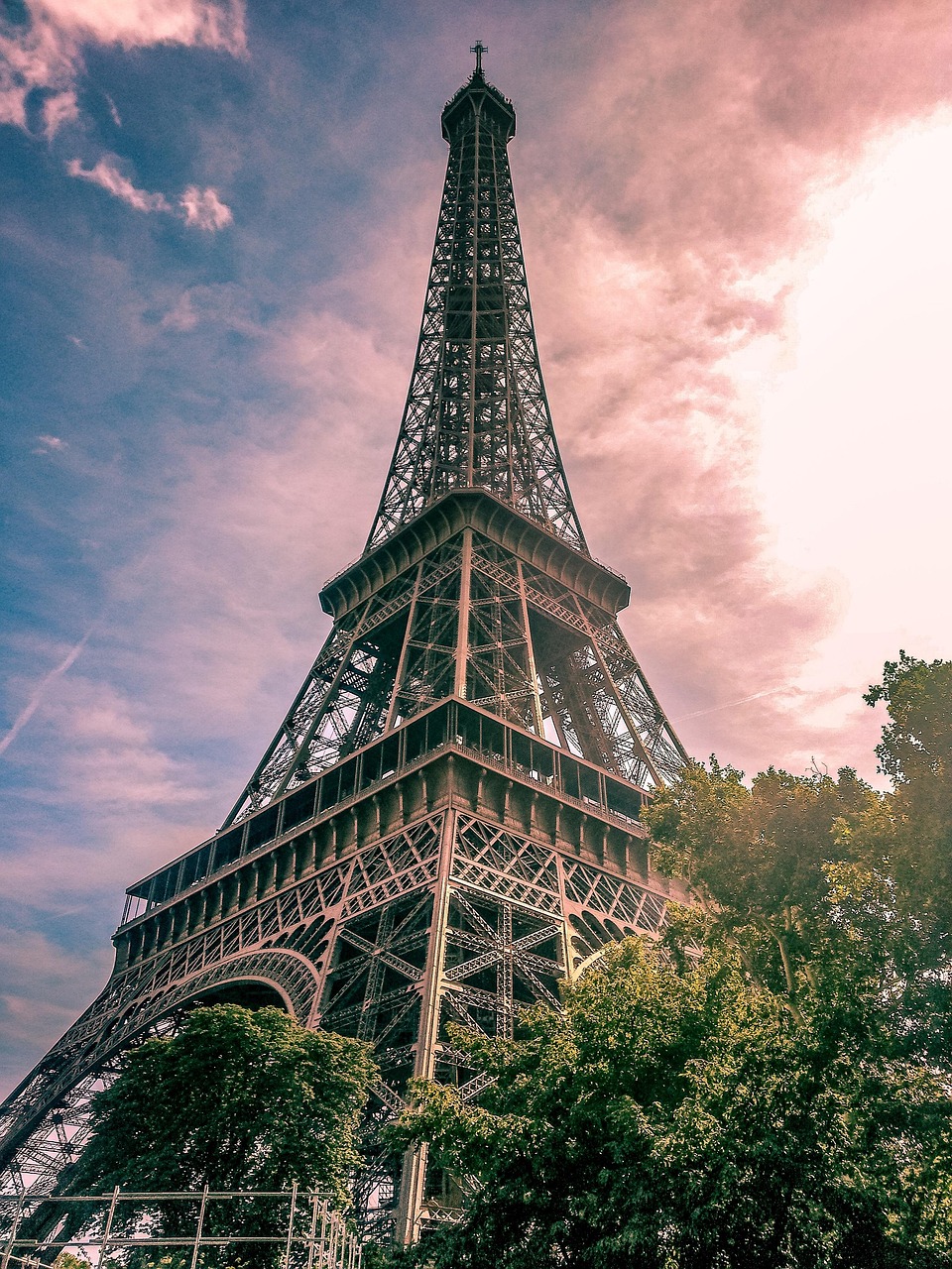Eiffel-torni kohoaa puiden ja pilvien keskellä, taustalla vaaleanpunainen ilta-aurinko ja sinertävä taivas. Eiffel-torni on Pariisin tunnetuimpia nähtävyyksiä