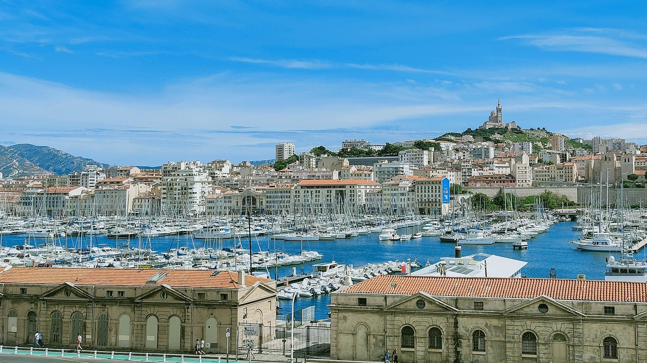 Marseille on satamakaupunki Ranskassa Satamalaiturit täynnä purjeveneitä, taustalla kukkulan kirkko ja sininen taivas.