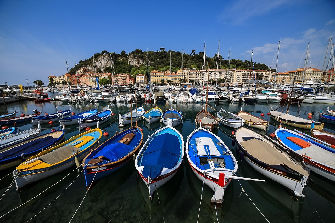 Nizza on Ranskan Riviera. Kaunis satamakaupunki. Sinisiä ja valkoisia veneitä satamassa, taustalla värikkäitä rakennuksia ja mäntymetsäinen kukkula.