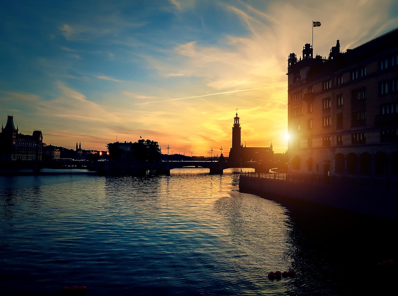 Tukholman kaupungissa on meri osa kaupunkia. Auringonlasku joen rannalla, historiallisia rakennuksia ja majakka, taivaalla pilviä ja heijastuksia vedessä.