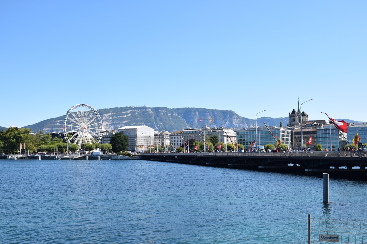 Geneve on suosittu matkakohde Sveitsissä. Rantamaisema Sveitsin kaupungista, suuri laiturisilta, Ferris-pyörä ja kirkontornit vuoritaustan edessä.