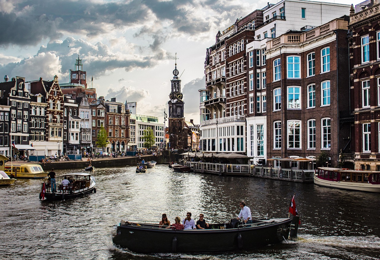 Amsterdam on Alankomaiden ja Hollanin eläväinen kanaalikaupunki. Kaupunkinäkymä kanavalla, tiilitaloja ja kirkontorni, veneitä ja turistit purjehtimassa pilvisellä säällä.