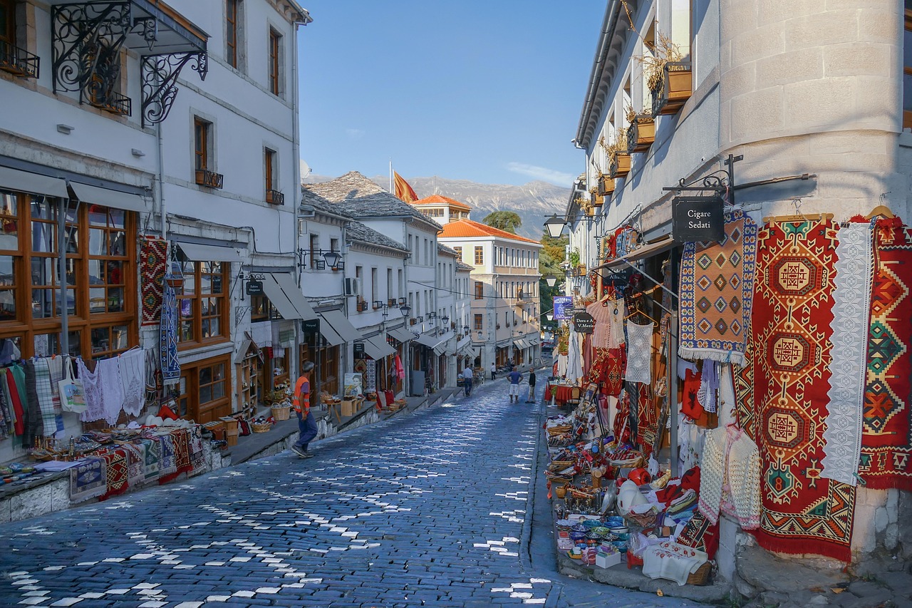 Gjirokastër, Albania on kaunis kaupunki Kaakkois-Albaniassa. Kapea mukulakivikatu, valkoisia rakennuksia ja värikkäitä mattoja myynnissä, taustalla vuoret ja sininen taivas.