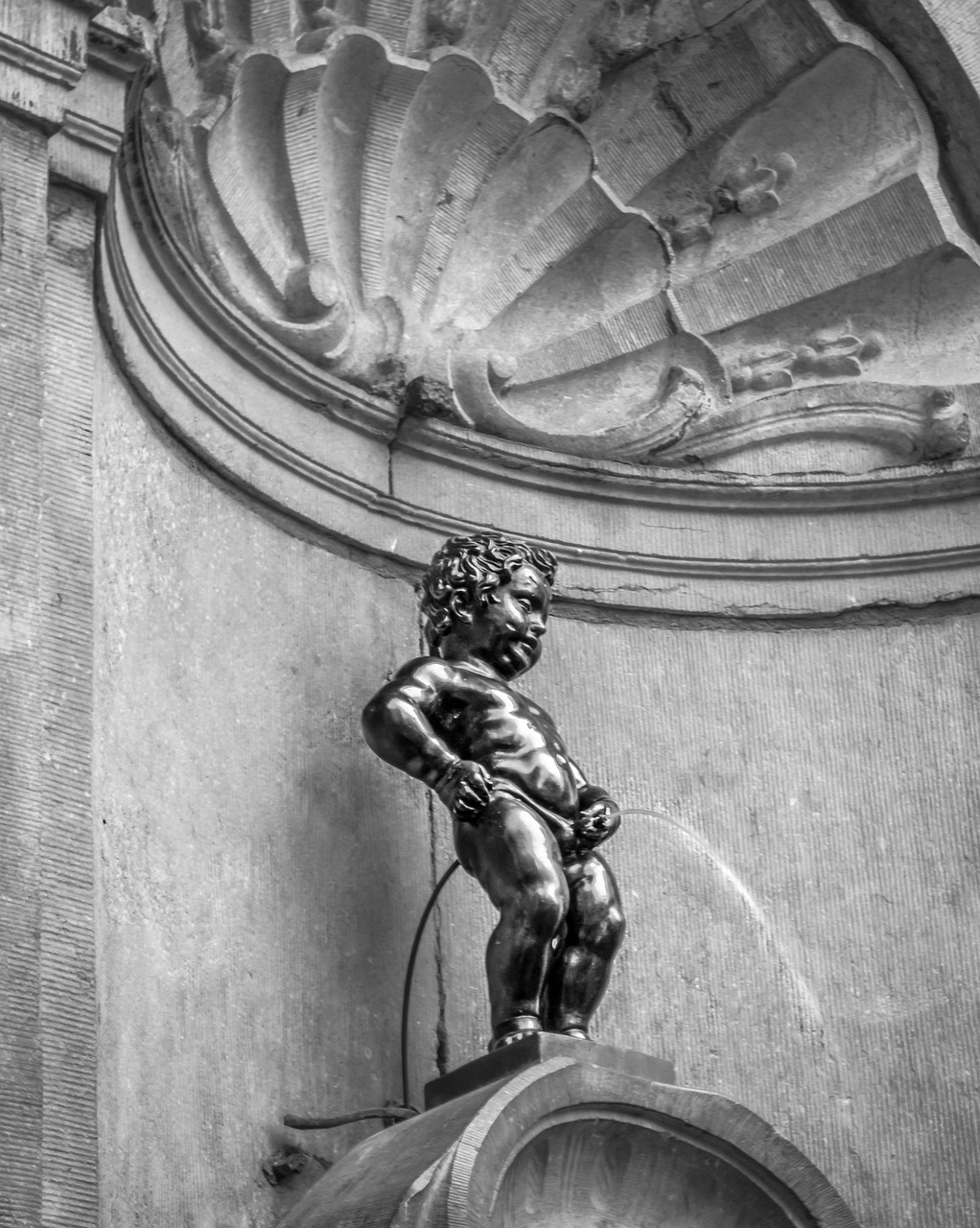 Belgia. Manneken pis on kuvatuin pronssipatsas Brysselin vanhassakaupungissa. Pronssinen putto-patsas seisoo jalustalla, taustalla koristeellinen kaari ja kohokuvioitu seinä.