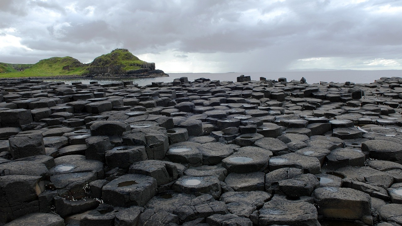 Giant's Causeway on asalttipylväistä koostuva nähtävyys Pohjois-Irlannin koillisrannikolla. Mustia basaltin kuusikulmaisia kivilohkareita rannalla, taustalla vihreä saari ja pilvinen meri.