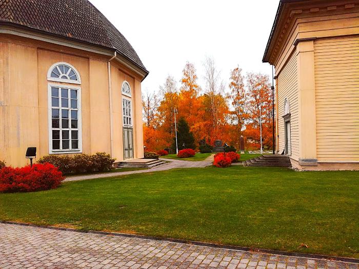 Vimpelin pyöreä kirkko on vihitty käyttöön 1811.
Kaksi vaaleaa rakennusta, siisti nurmikko ja punaiset pensaat, taustalla syksyiset puut oransseina.