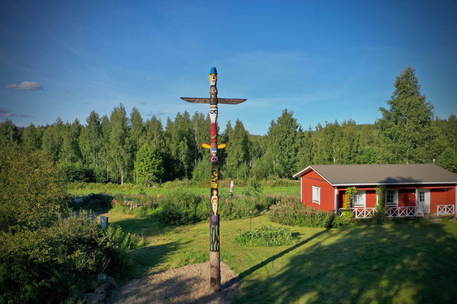 Vesannolla on pystyssä 21 huikeaa Sydänmaanpaalua, jotka jokainen kertovat juttuja Vesannon kunnasta.
Värikäs puinen totemipylväs seisoo punaisen mökin edessä, taustalla koivumetsä ja sininen taivas.