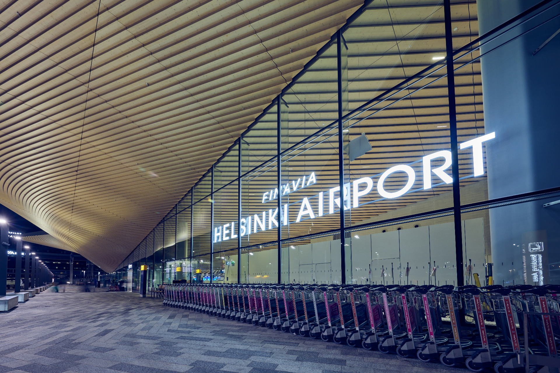 Helsinki-Vantaan lentokenttä on Suomen portti maailmalle.
Pitkän käytävän katto ja lasiseinät, neonvalaistu teksti sekä rivissä pysäköityjä polkupyöriä.