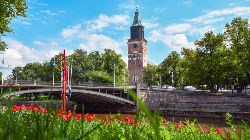 Turun tuomiokirkko on Arkkipiispan ja Turun piispan kotikirkko.
Punatiilinen kirkontorni kohoaa sillan takaa, ympärillä vehreää puistoa ja punaisia kukkia sinisellä taivaalla.