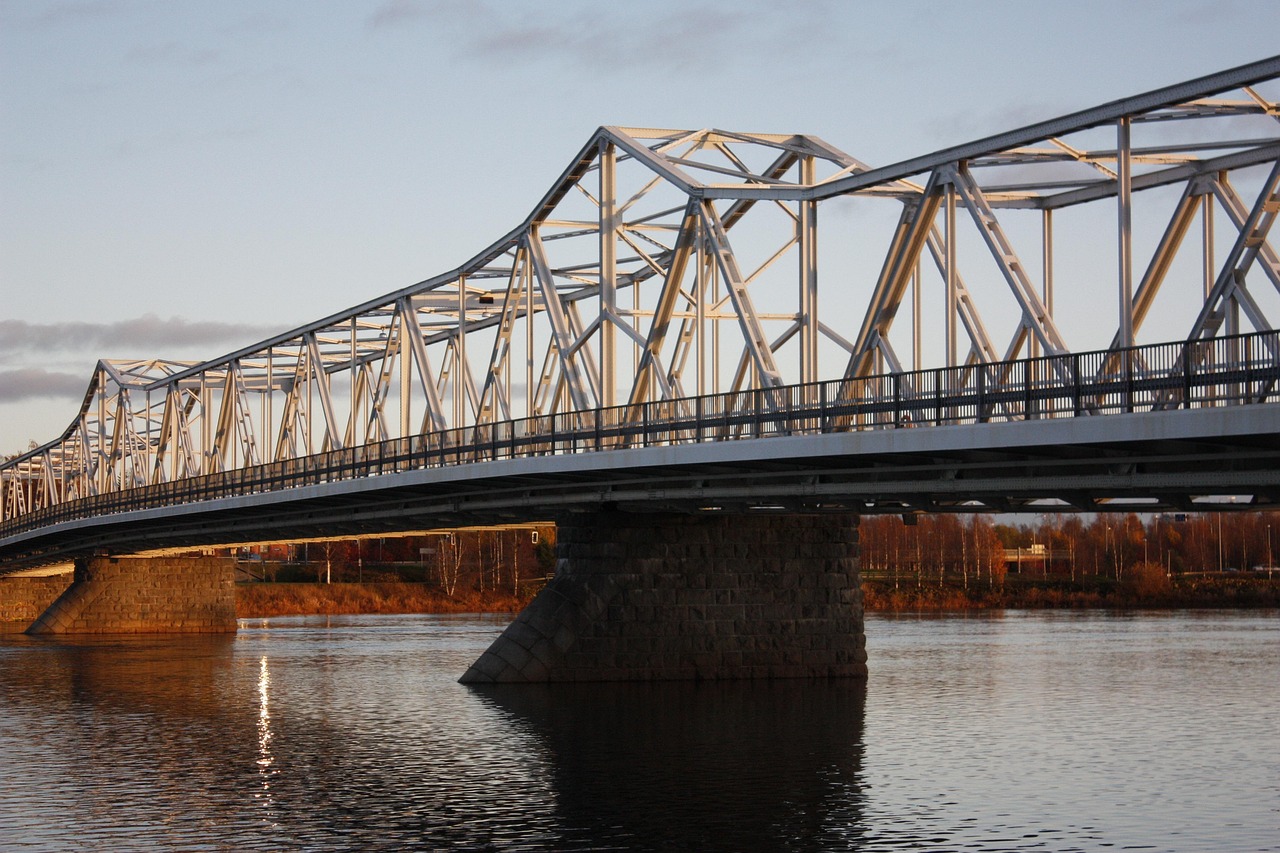 Hannulan silta. Torniojoki.
Teräksinen rautatiesilta kaartuu joen yli, näkyy ristikkorakenne ja betoninen tukimuuri, ilta-auringon valossa.