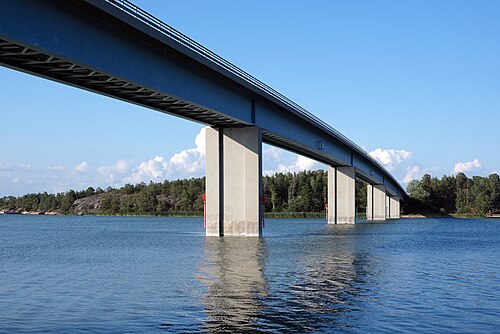 Kaitaisten silta, Taivassalossa on 477 metriä pitkä.
Silta ylittää järven, betonipilarit ja teräksinen kaari näkyvät veden heijastuksissa sinisellä taivaalla.