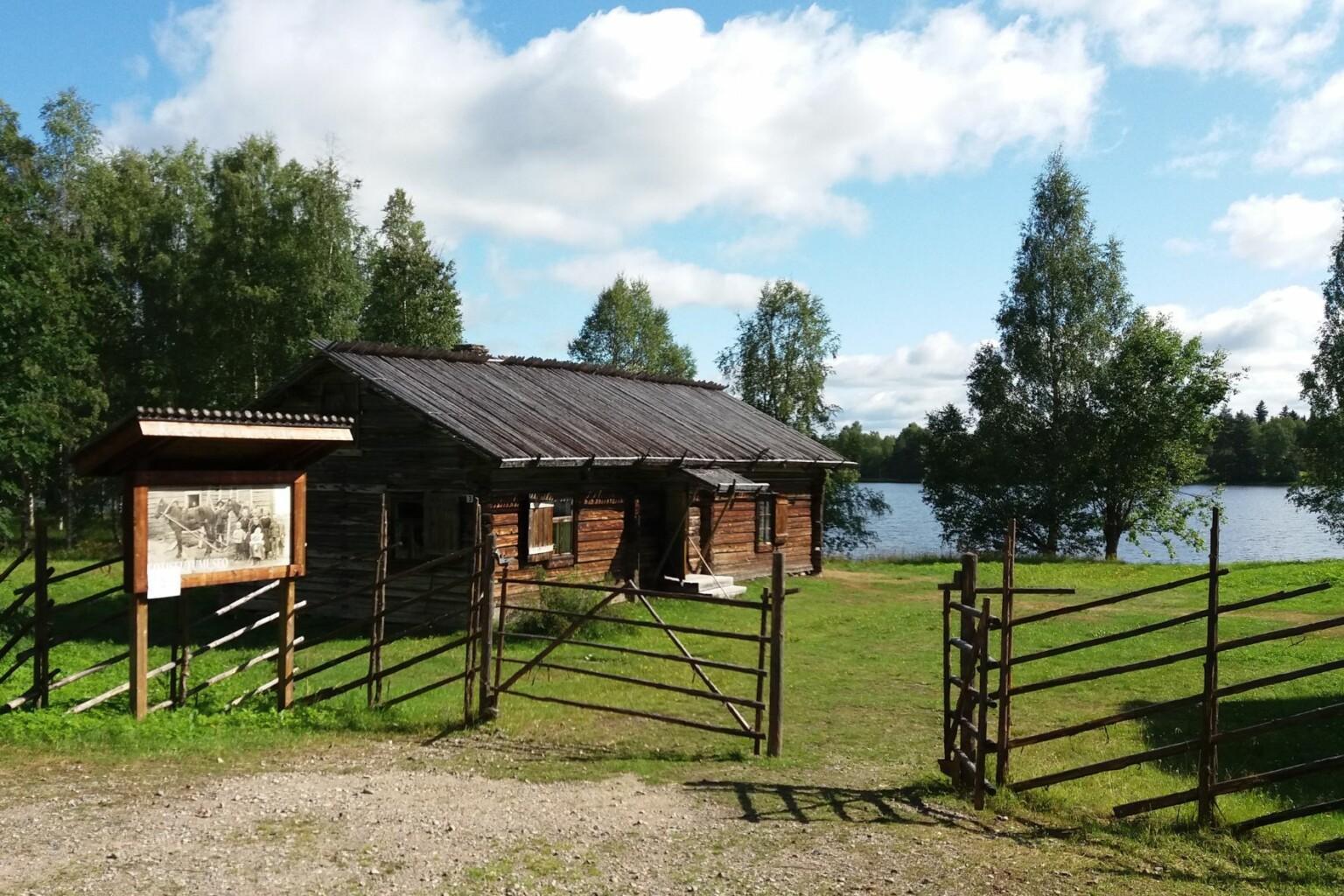 Taivalkosken kotiseutumuseo, Taivalkoski.
Vanha hirsinen rakennus järven rannalla, aita ja vihreä nurmikko, taustalla pilvinen sinitaivas.