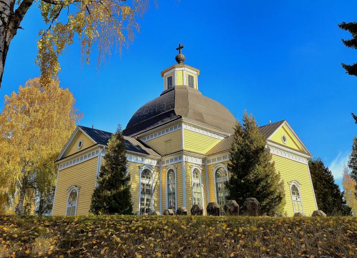 Taipalsaaren puinen kirkko on valmistunut 1754.
Keltainen puukirkko, kupolikellotorni ja kaarevat ikkunat, syksyinen lehtimaisema sinistä taivasta vasten.